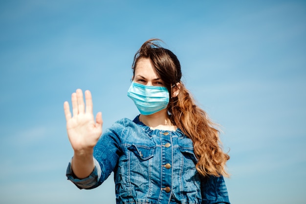 Photo woman in protective sterile medical mask on her face looking at camera outdoors. hand stop sign . pandemic coronavirus concept.