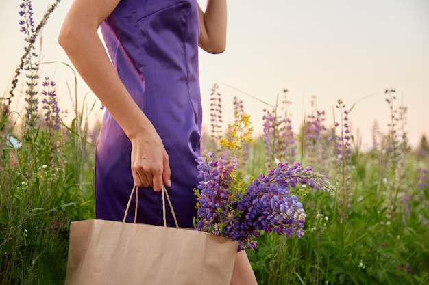 Woman in purple dress with craft bag of lupinus in the meadow Lupin lupine field with purple flowers at sunset Natural products shopping concept Wellness and natural concept