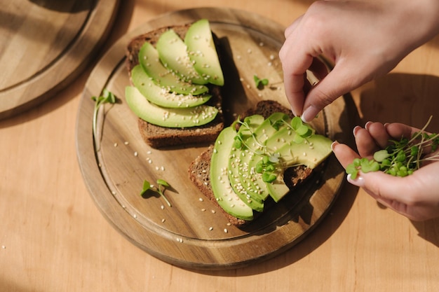 Woman put mustard seedlings on avocado sandwich with dark rye bread made with fresh sliced avocados from above with sesame seeds