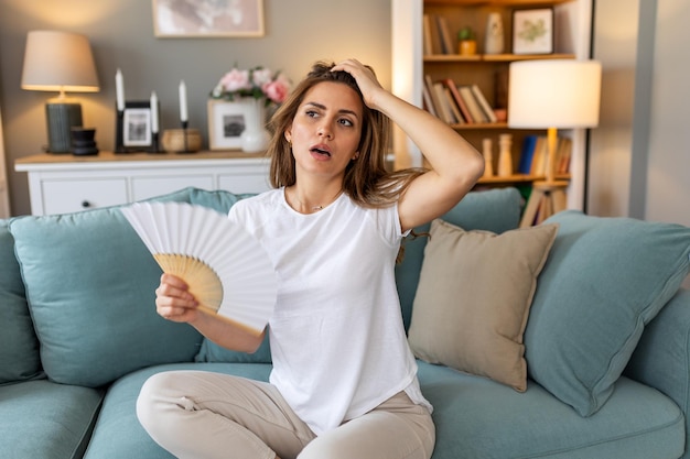 女性はソファのクッションに頭を置き耐え難い熱波のためゆっくりと感じます手のファンで自分自身を冷却します 暑い夏エアコンなしのアパート気候制御システムのコンセプト