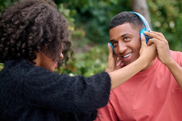 Woman putting on headphones on smiling man outdoors