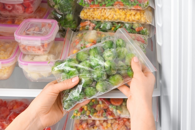 Woman putting plastic bag with frozen vegetables into refrigerator