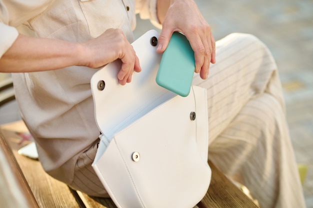 Woman putting smartphone in white handbag