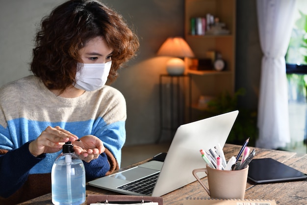 Photo woman in quarantine and social distancing wearing surgical mask and cleaning hands with alcohol gel sanitizer while working from home during covid-19 coronavirus pandemic