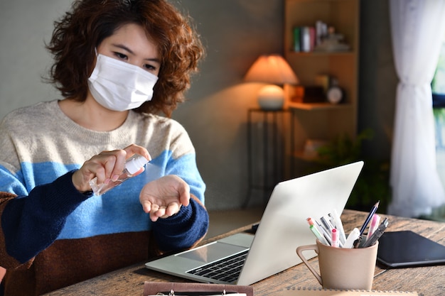 Foto donna in quarantena e distanza sociale che indossa una maschera chirurgica e si pulisce le mani con disinfettante in gel alcolico mentre lavora da casa durante la pandemia di coronavirus covid-19