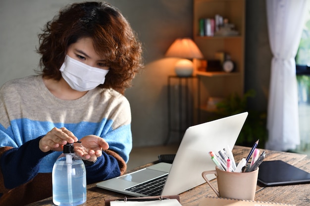 Photo woman in quarantine and social distancing wearing surgical mask and cleaning hands with alcohol gel sanitizer while working from home during covid-19 coronavirus pandemic