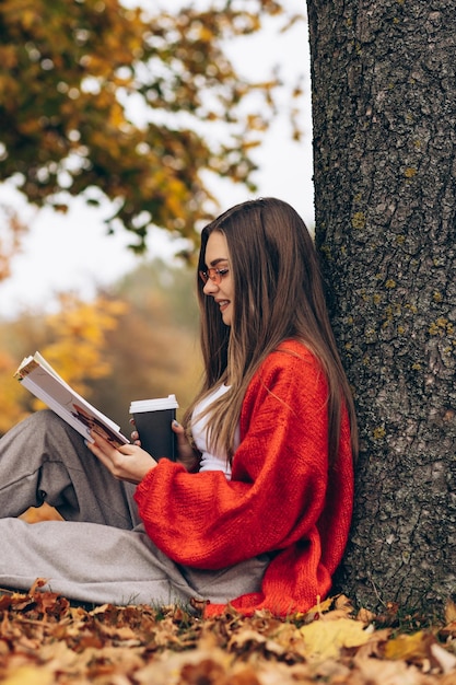 秋の公園で読書をし、木の下でコーヒーを飲む女性