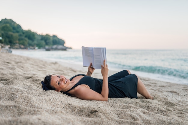 Donna che legge un libro sulla spiaggia. guardando la fotocamera