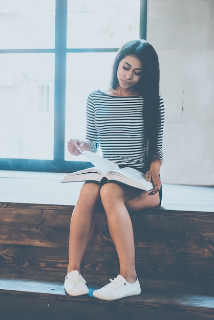 Photo woman reading book. beautiful young mixed race woman reading a book while sitting indoors and near the large window