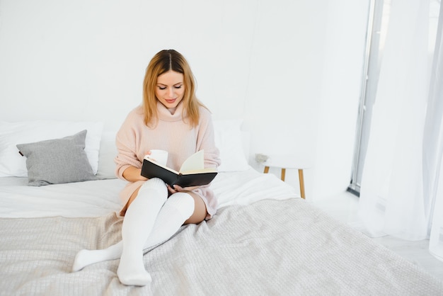 Una donna che legge un libro e sorride mentre si siede a letto. la sveglia sulla scrivania accanto a lei.