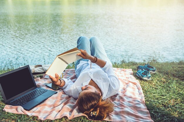 Photo woman reading book while lying by laptop at lakeshore