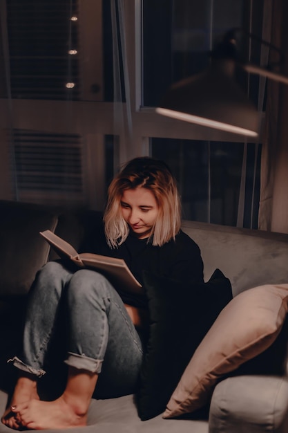 Женщина читает книгу, сидя дома на диване.