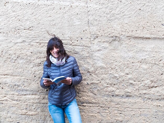 Woman reading book while standing against wall