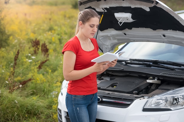 Woman reading owner manual at broken car in meadow