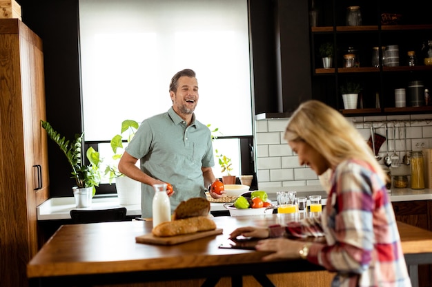 A woman reads from a tablet at the kitchen counter while a man holding an tomatoes smiles at her