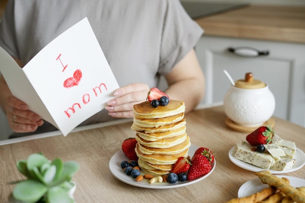 女性が子供から母の日のギフトカードを読む朝食のサプライズパンケーキとイチゴとブルーベリー