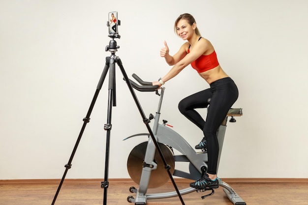 Woman recording tutorial video on cell phone on tripod riding exercise bike and showing thumb up