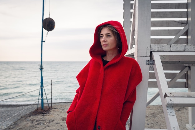 Foto una donna in cappotto rosso sta in riva al mare, appoggiata a una struttura in legno.