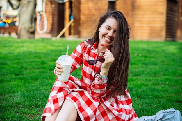 Woman in red dress drinking smoothie while sitting on green grass in park