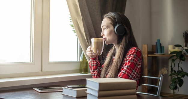 A woman in a red dress is listening audiobook with headphones and drinking coffee by the window Concept of modern technological education and the study of books with joy