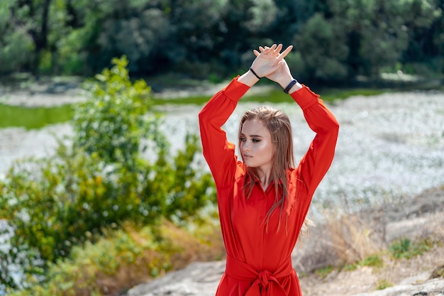 Photo a woman in a red dress is standing on a rocky shoreline with her arms raised in the air the scene is serene and peaceful