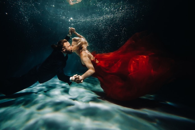 A woman in a red dress and a man in a suit are kissing underwater.A pair of floats under water.