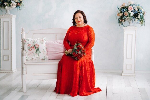 A woman in a red dress sits on a sofa and holds a bouquet of red roses and strawberries in the interior.