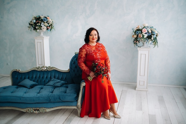 A woman in a red dress sits on a sofa and holds a bouquet of red roses and strawberries in the interior.