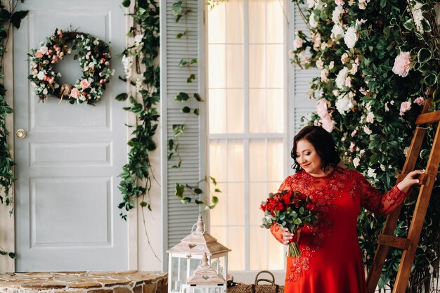 A woman in a red dress stands and holds a bouquet of red roses and strawberries in the interior.