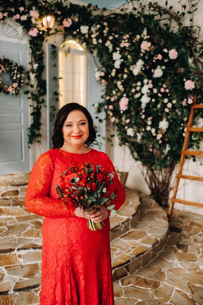 A woman in a red dress stands and holds a bouquet of red roses and strawberries in the interior.