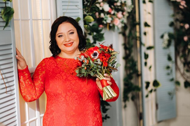 Женщина в красном платье стоит и держит в интерьере букет красных роз и клубники.