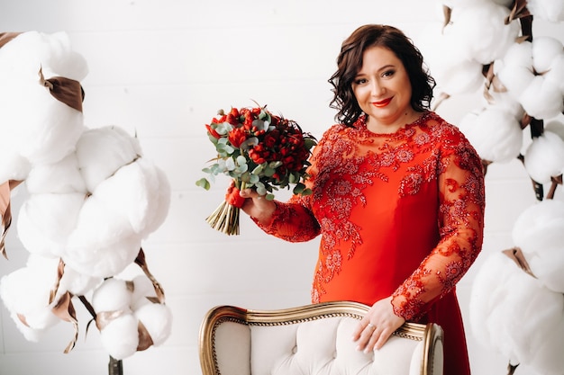 A woman in a red dress stands and holds a bouquet of red roses and strawberries in the interior.