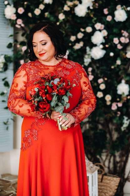 A woman in a red dress stands and holds a bouquet of red roses and strawberries in the interior.