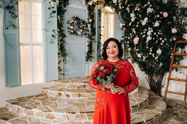 A woman in a red dress stands and holds a bouquet of red roses and strawberries in the interior