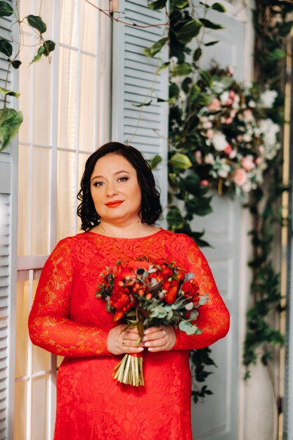 A woman in a red dress stands and holds a bouquet of red roses and strawberries in the interior.