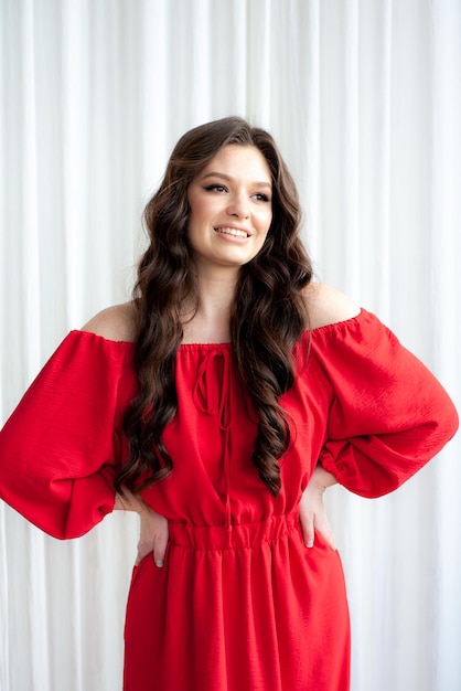 A woman in a red dress with long curly hair stands in front of a white curtain.