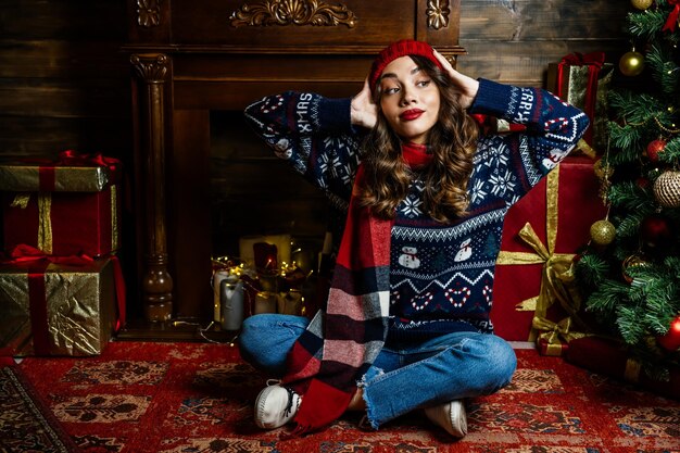 A woman in a red hat and scarf holds her head A beautiful brunette sits under a Christmas tree against the backdrop of New Year's decorations