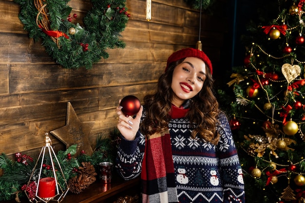 赤い帽子とスカーフをかぶった女性が喜ぶ 美しいブルネットがクリスマスツリーとクリスマスの飾りを背景に暖炉のそばに立つ