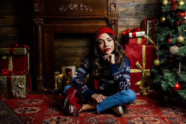 A woman in a red hat and scarf thought A beautiful brunette sits under a Christmas tree against the backdrop of New Year's decorations