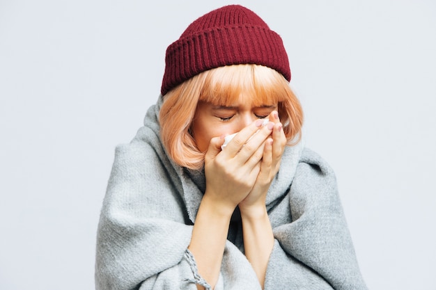 Woman in red hat, warm scarf with paper napkin sneezing, experiences allergy symptoms, caught a cold, closed eyes