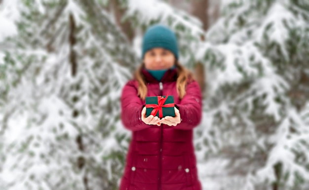 woman in red holding in hands Gift green box with red ribbon against snowy tree in winter Christmas