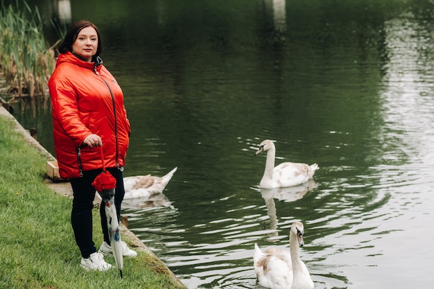 赤い上着を着て傘をさしている女性が白鳥のいる湖の近くの夏の公園を歩く