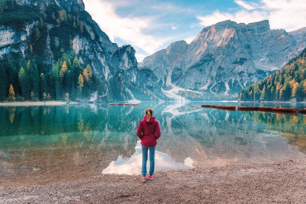 Woman in red jacket is standing on the coast of Braies lake at sunrise in autumn. Dolomites, Italy.