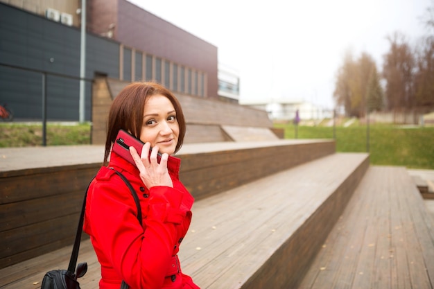 Una donna con un impermeabile rosso sta parlando al telefono vicino a un business center. una bruna con un taglio di capelli a caschetto in autunno o in primavera conduce affari, comunica a distanza