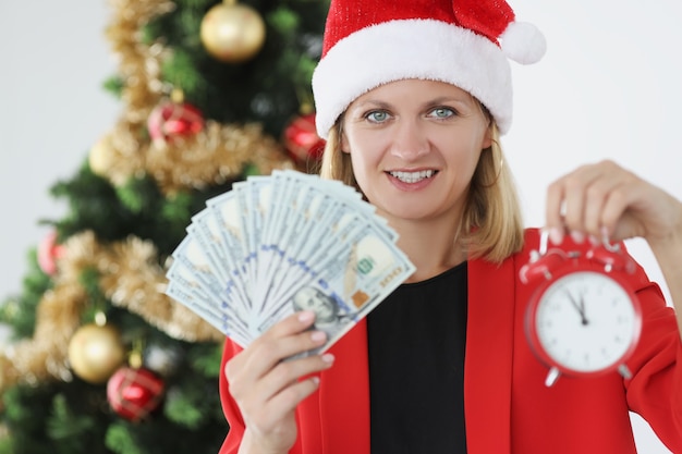 새 해 나무 근처 손에 돈과 알람 시계를 들고 빨간 산타 모자에 여자. 새해 개념의 성공적인 비즈니스를 위한 전략