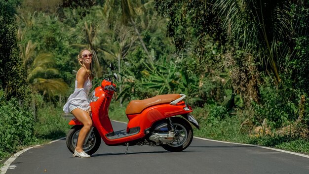 Woman on red scooter in white clothes drive on forest road\
trail. dancing trip. one girl caucasian tourist in sunglasses\
dance, relax, rest near motorbike. asia thailand ride tourism.\
motorcycle rent.