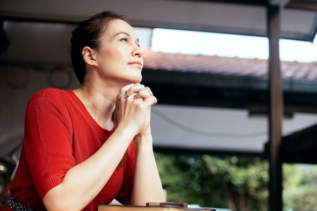 Photo a woman in a red shirt sits at a table with her hands folded in prayer.