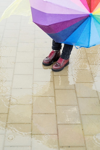 Foto donna con le scarpe rosse che tiene l'ombrello arcobaleno fuori nella pioggia in piedi in una pozzanghera