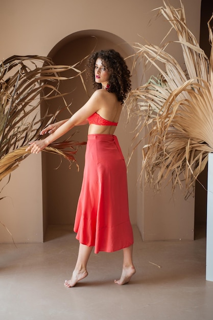 A woman in a red skirt stands in a room with a palm plant in the background