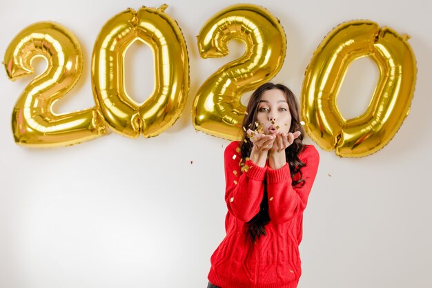 Foto donna in maglione rosso che soffia coriandoli d'argento davanti al 2020 palloncini di capodanno